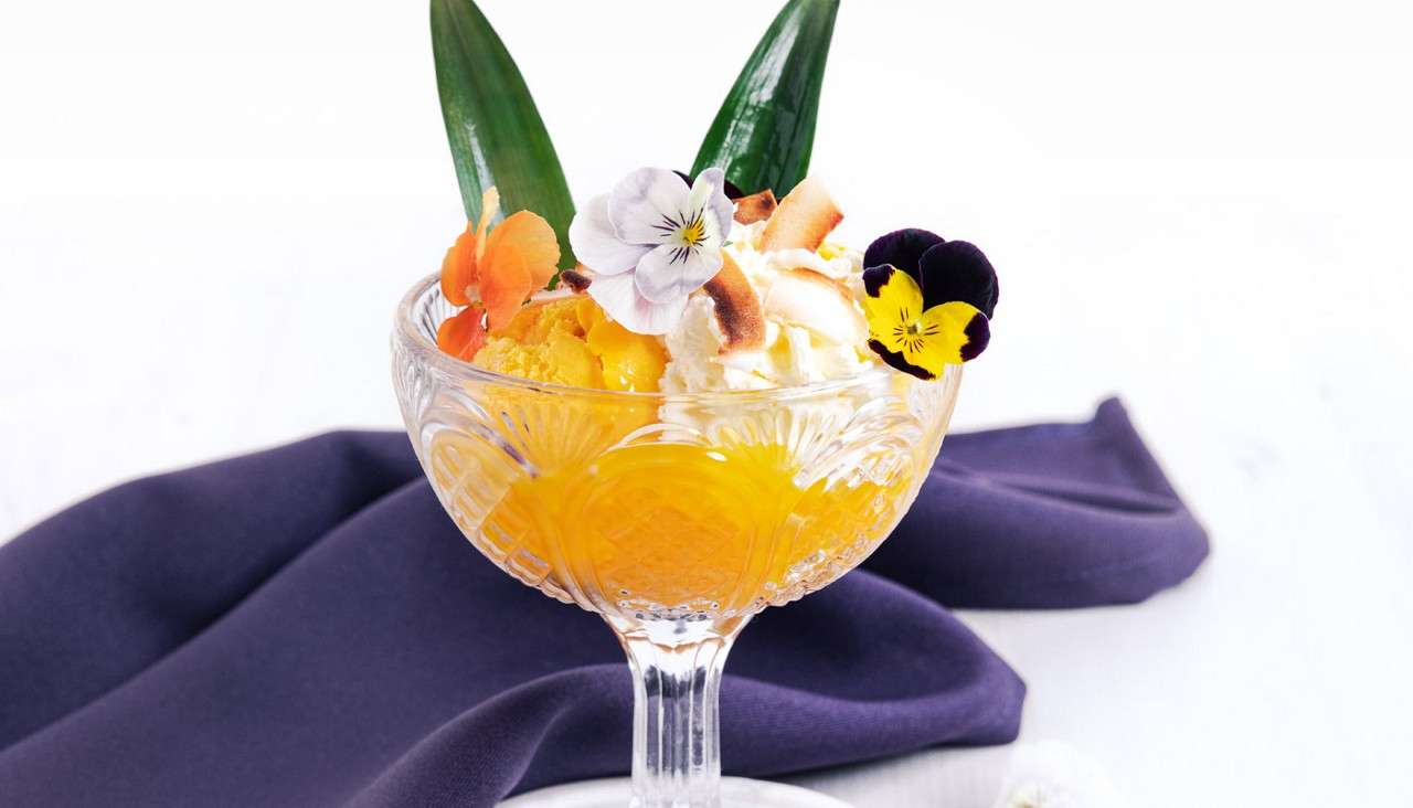 Crystal coupe dessert holds orange sorbet and whipped cream, topped with toasted coconut, pansy and orange blossoms, and two green leaves; resting on a purple napkin against a white background.