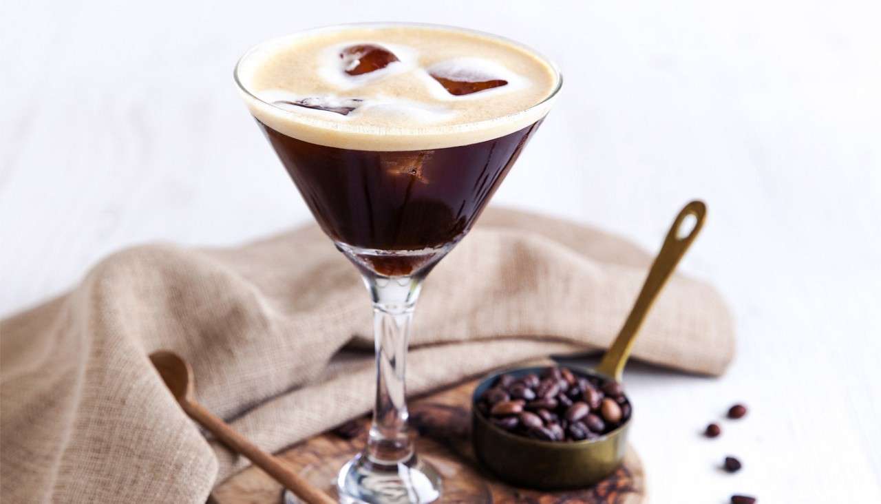 Martini glass holds dark iced coffee topped with foam, resting on a wooden board, accompanied by a brass scoop of coffee beans and a folded beige cloth against a bright white background.