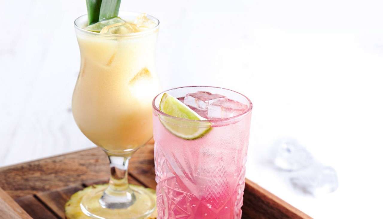 Two chilled cocktails sit together: a creamy yellow drink with ice and pineapple leaf, and a pink iced drink with lime wedge, on a wooden tray against a white background.