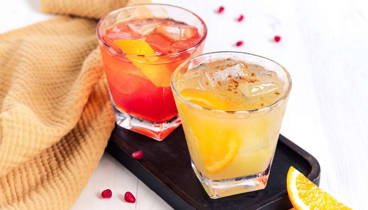 Two cocktail glasses on a black tray: one yellow with orange slices and cinnamon, one red with ice and orange peel; beige cloth, pomegranate seeds, and an orange wedge nearby.