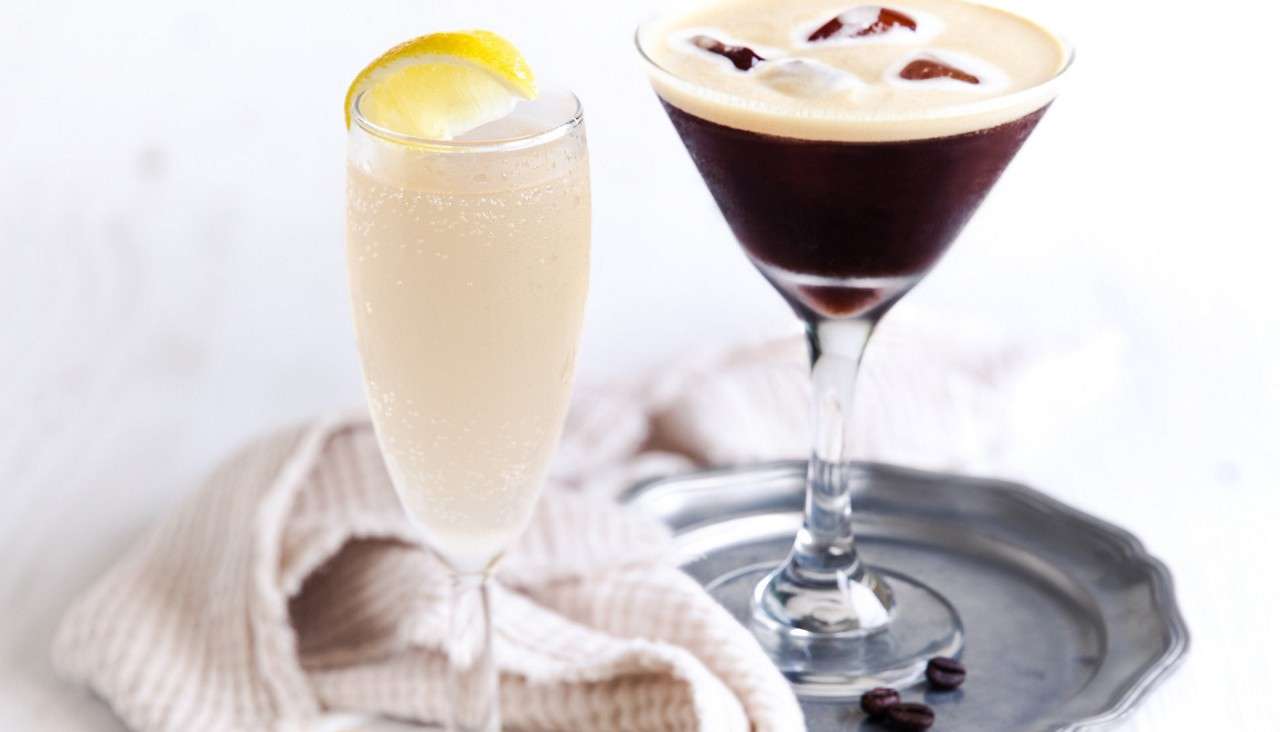 Two cocktails rest side by side: a pale, bubbly flute with a lemon wedge, and a dark, frothy martini in a stemmed glass on a metal tray, coffee beans nearby.