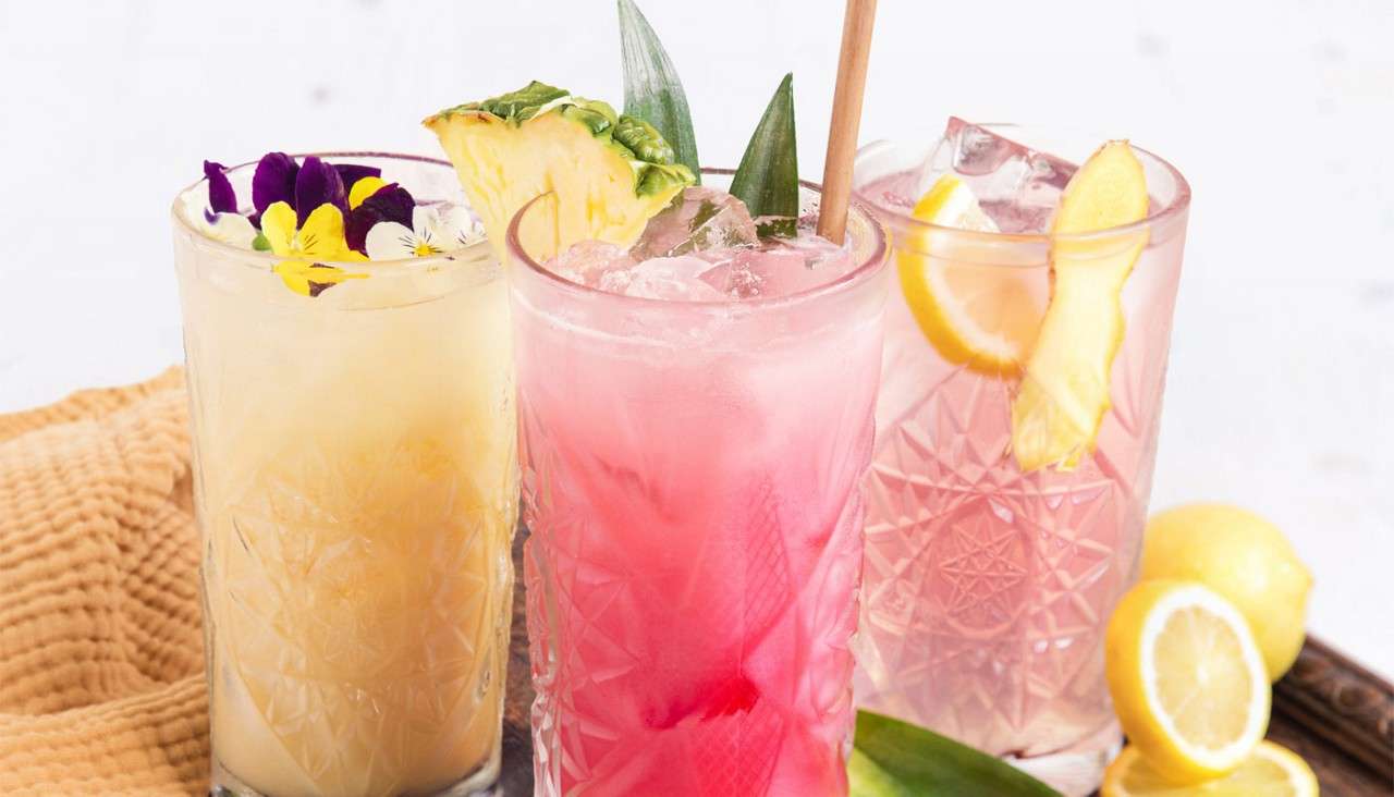 Three iced beverages sit in patterned glasses, garnished with pineapple, edible flowers, and lemon slices; they rest on a table with citrus and fruit pieces, against a bright, minimal background.