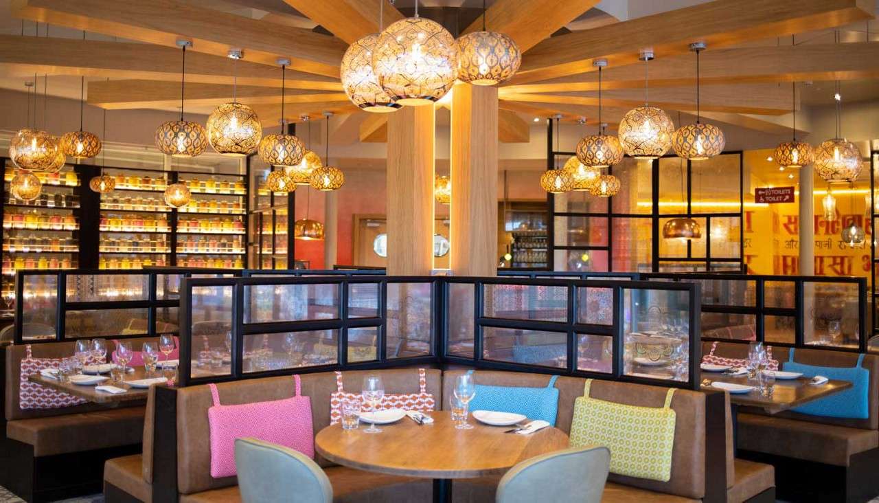 Round dining table is set with plates and glasses, surrounded by colorful cushioned booths; ornate pendant lights glow overhead; background shows jars on shelves and red sign reading "TOILETS →".