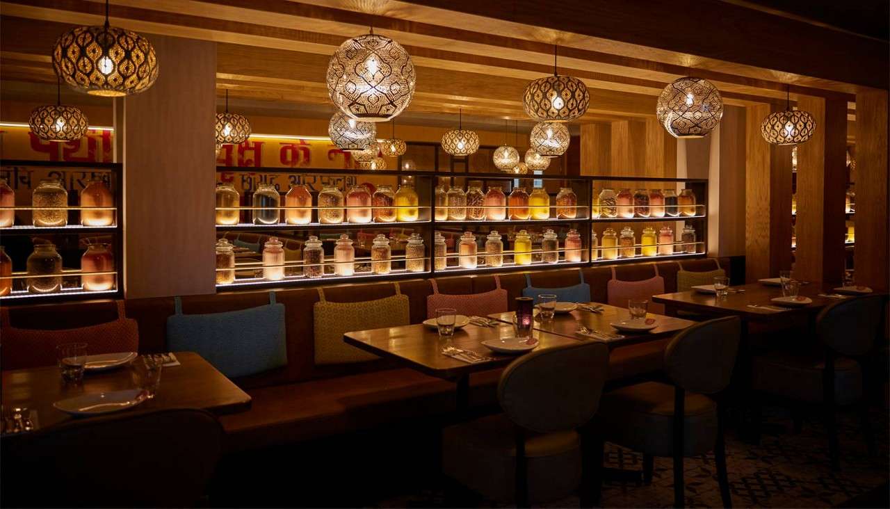 Shelved glass jars glow under strip lights; pendant lamps illuminate set dining tables and cushioned booths in a warm wooden restaurant. Text: [Hindi script on back wall, partially obscured/unreadable].