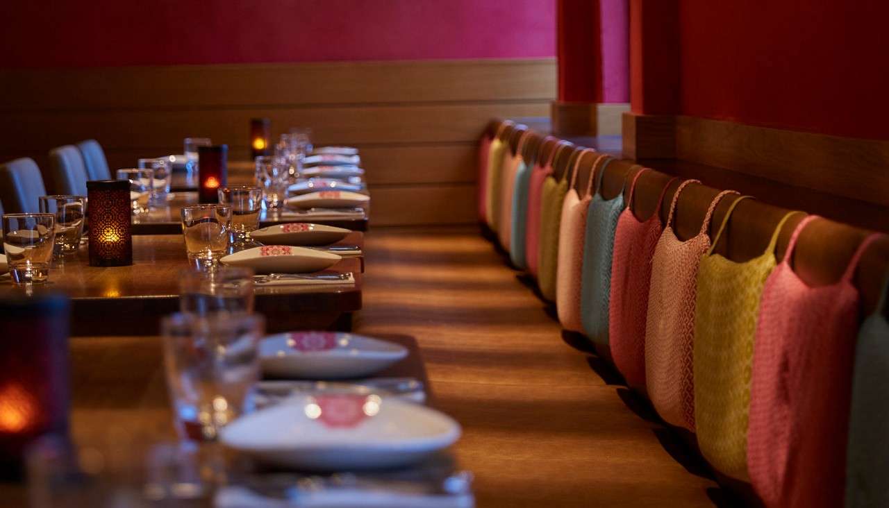 Long dining table sits prepared with glassware, cutlery, and folded napkins; votive candles glow. Context: a modern restaurant featuring a bench lined with colorful textured backrest covers against red walls.