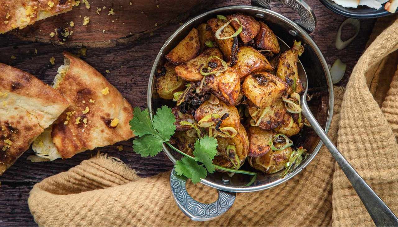 Roasted potato wedges sit in a metal bowl, garnished with scallions and cilantro. A spoon rests inside, beside torn naan and a mustard-colored towel on a rustic wooden table.