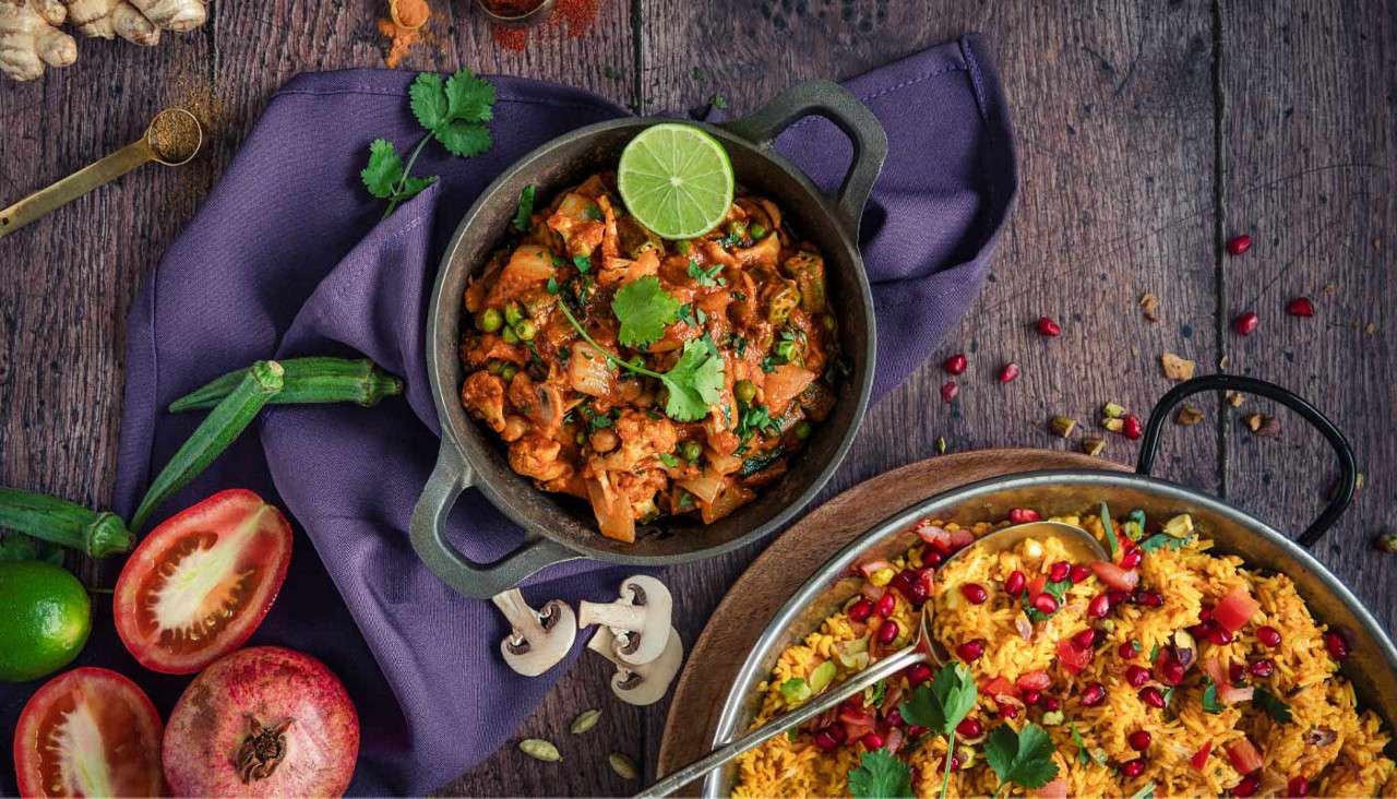 Two Indian dishes sit ready: vegetable curry in a cast-iron pot and yellow rice with pomegranate in a metal pan, garnished with cilantro, surrounded by tomatoes, okra, limes, spices, mushrooms.