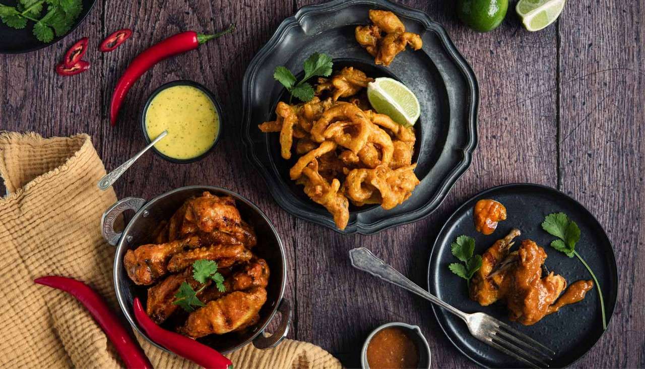 Fried chicken wings sit sauced and arranged; crispy fritters rest with lime wedges; bowls of yellow and brown sauces, red chilies, and cilantro garnish on a rustic wooden table.