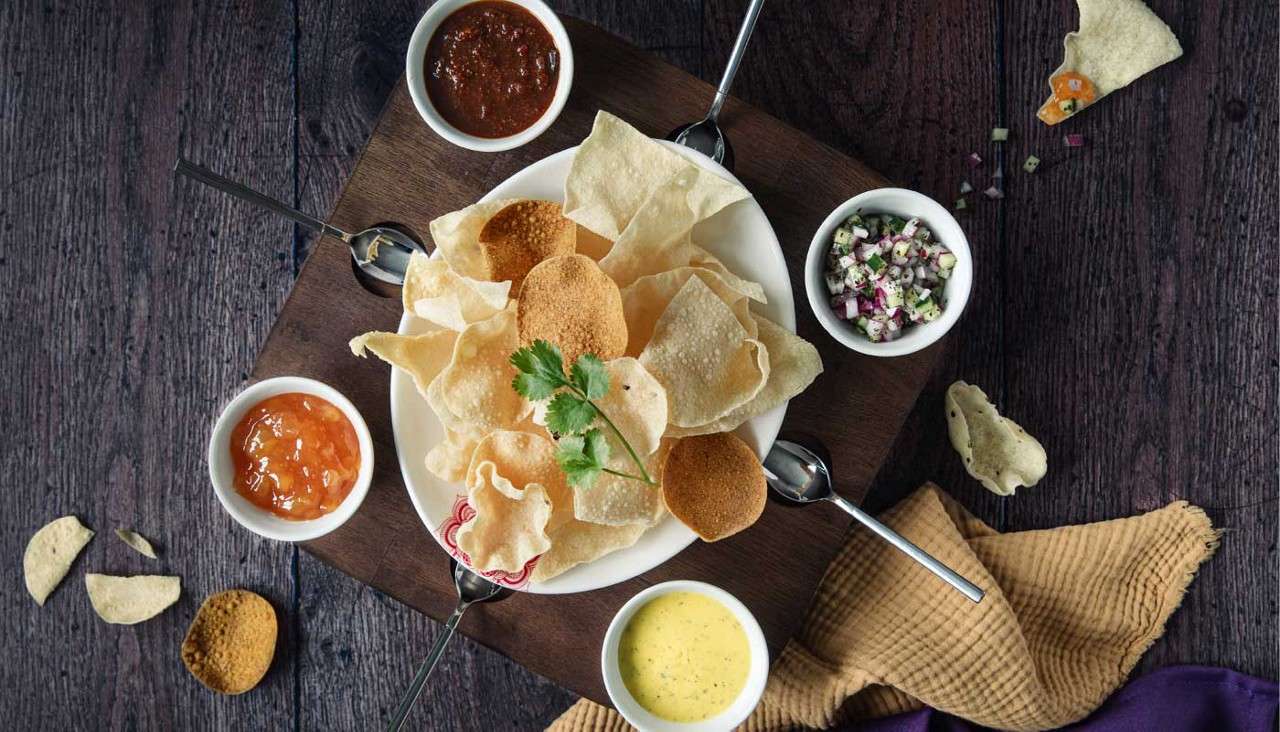 Crispy papadum chips sit in a white bowl, topped with cilantro, served with four small bowls of chutneys and diced salad, arranged on a board over a dark wooden table.