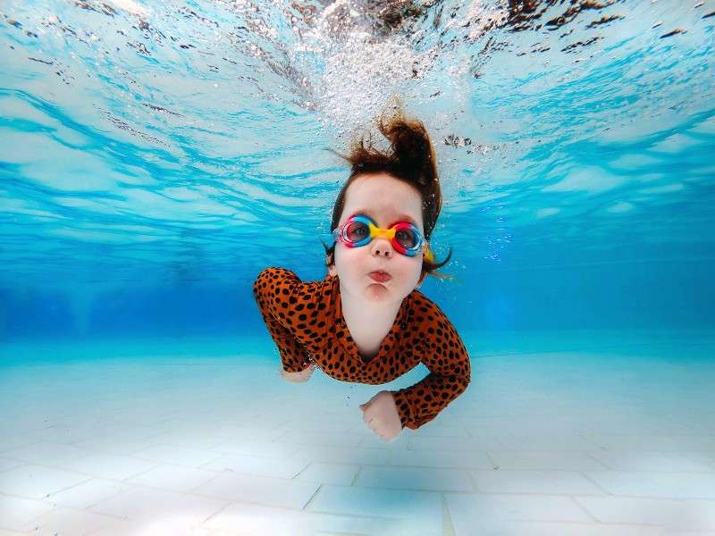 Child in colorful goggles swims underwater, cheeks puffed, wearing orange black-spotted swimsuit; arms bent, hair floating upward, in a clear blue swimming pool with tiled floor and rippling surface above.