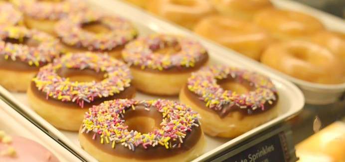 Frosted ring donuts rest on a bakery tray, topped with chocolate glaze and rainbow sprinkles, with plain glazed donuts stacked behind them in a lit glass display case.
