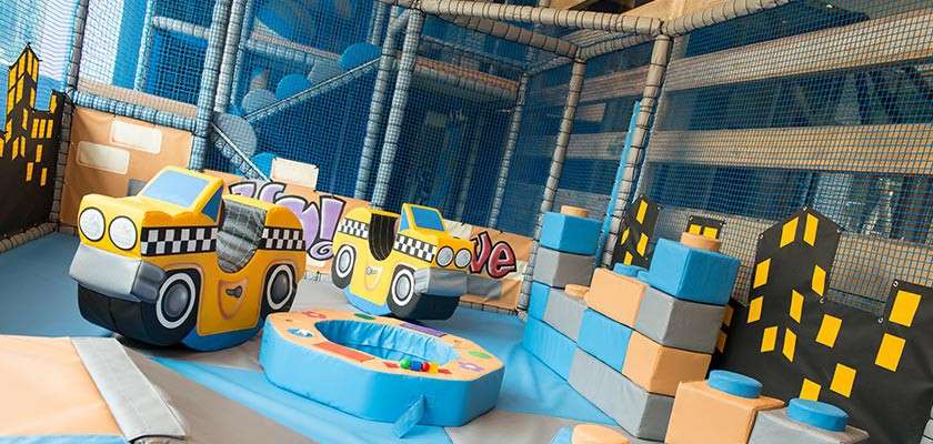 Two foam taxi cars sit side-by-side, inviting play, amid padded blocks and a small circular ball pit, inside a netted indoor soft-play cityscape; graffiti-style text reads: love.