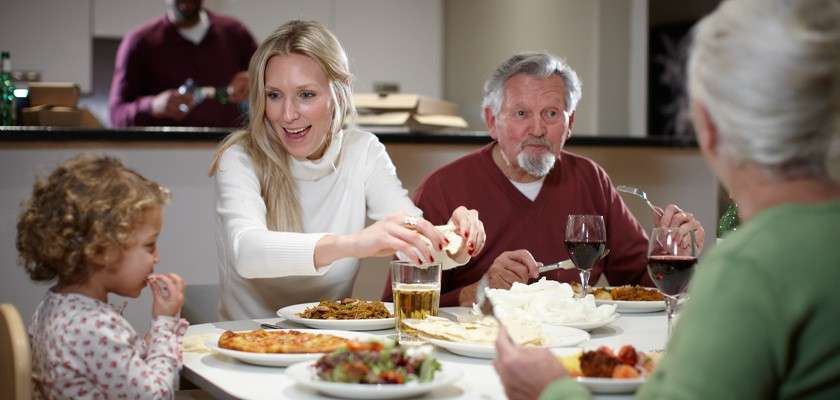 Family members share dinner, passing flatbread and eating, around a white table. Plates of salad, pizza, curries, and drinks sit before them, while a kitchen counter and person stand behind.