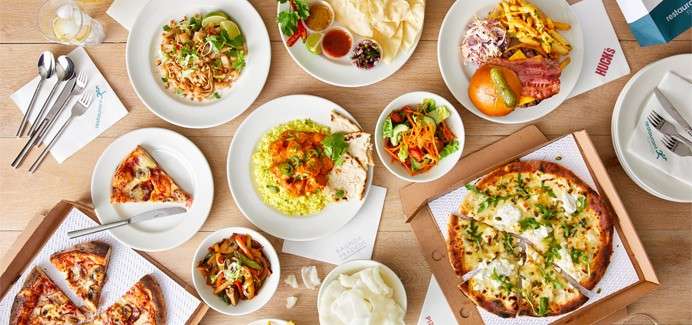 Assorted dishes sit arranged on a wooden table: two pizzas in boxes, pasta, curry with rice, naan and dips, fries with meat, salads, vegetables, cutlery and drinks, prepared for sharing.