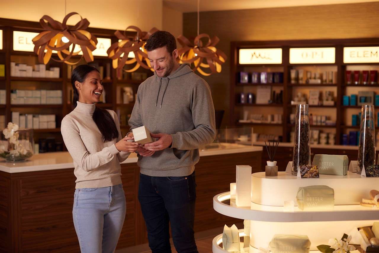 Two people examine a small boxed product, standing in a warmly lit beauty store with skincare shelves and displays. Visible text: OPI; A BETTER WAY.