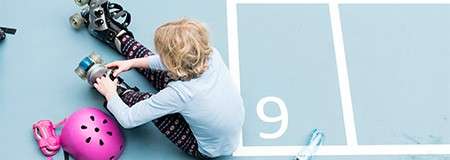 Person tightens roller skates while seated on a blue court; a pink helmet and pads lie nearby; white court marking reads “9.”