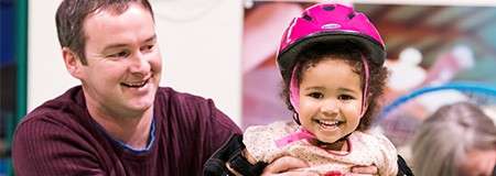 Child wearing a pink helmet smiles while being steadied by an adult man; indoors, possibly at a play area, with blurred background and others nearby.