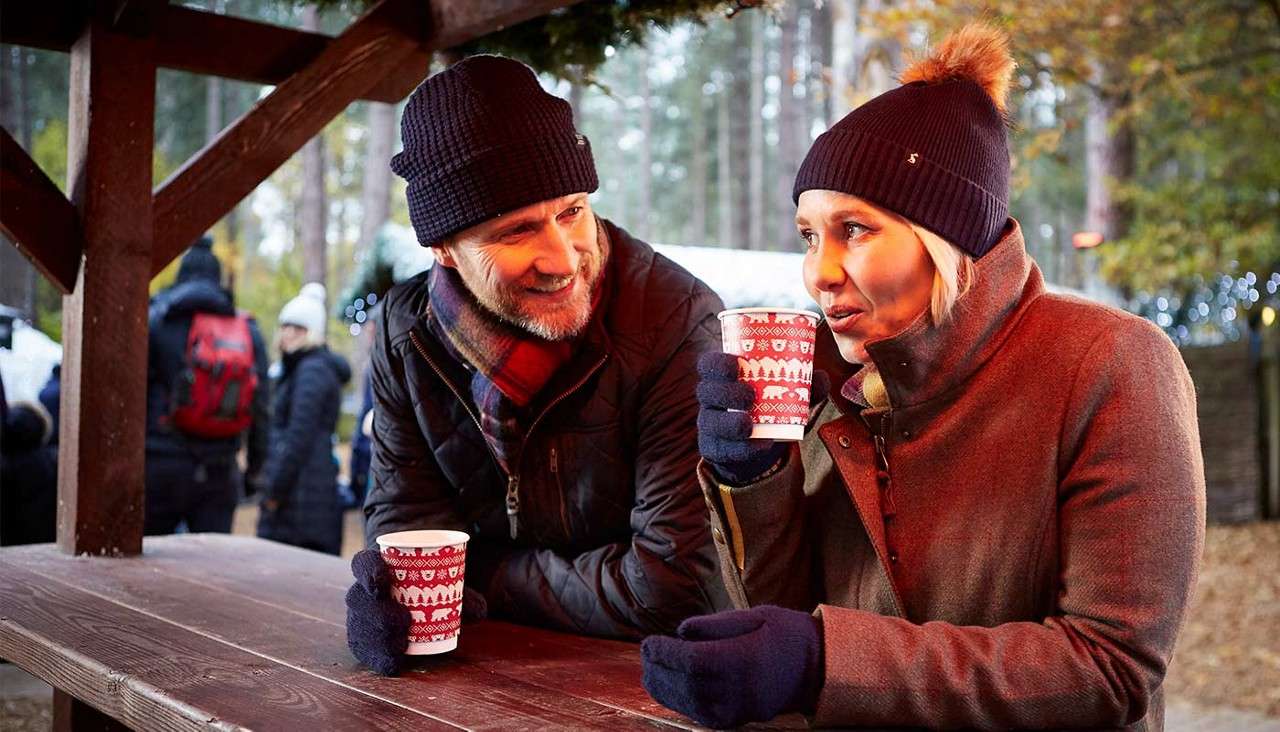 Two bundled adults drink hot beverages from red patterned cups, chatting at a wooden table in an outdoor winter market or park, with other visitors and string lights among trees.
