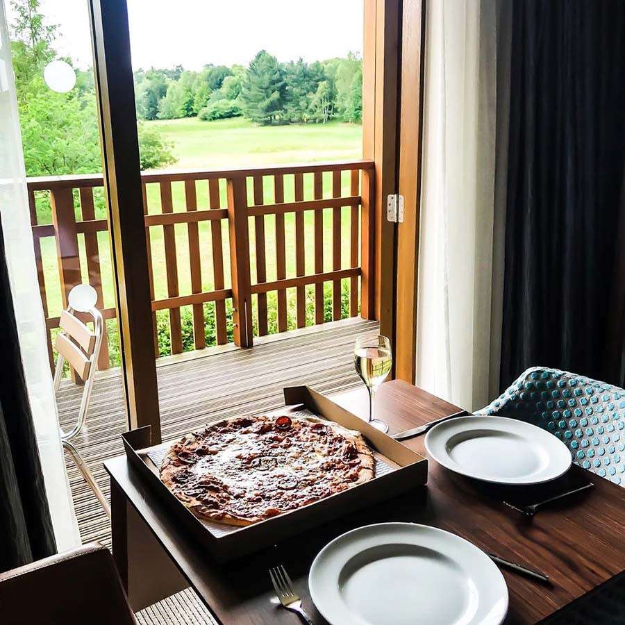 Pizza rests in an open box on a wooden table with two plates and a wine glass, beside an open balcony door overlooking a grassy field and trees.