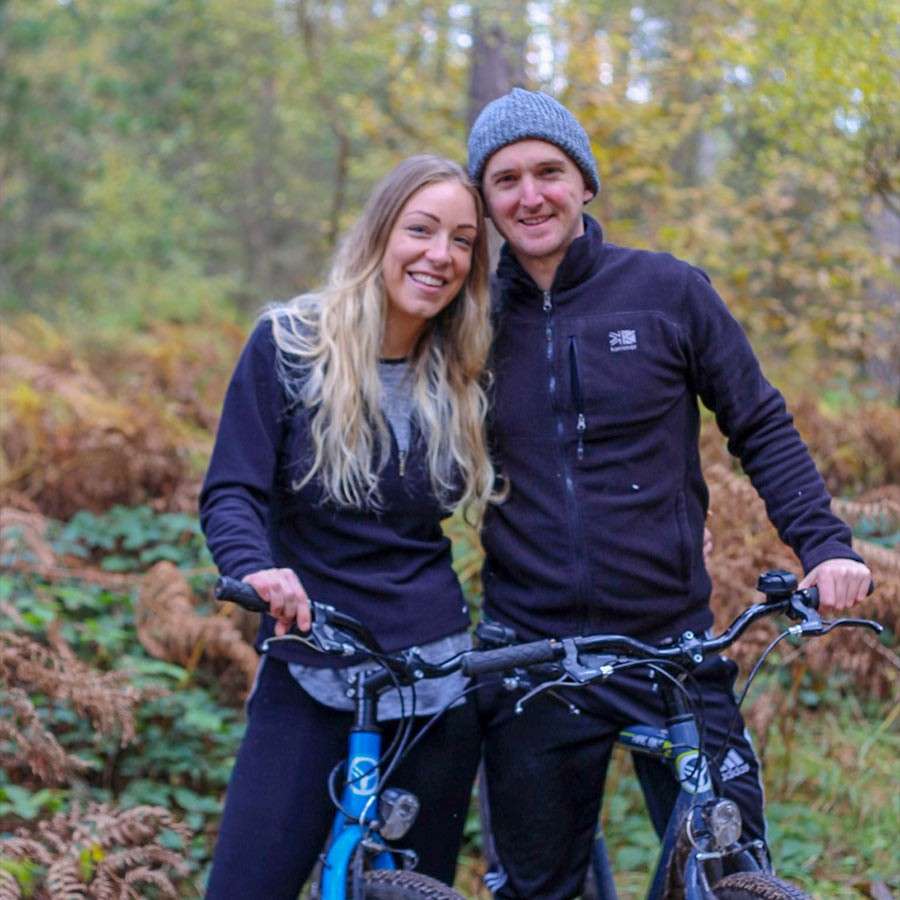 Two people stand, smiling, holding mountain bikes, pausing on a leafy forest trail with autumn ferns and trees; dressed in dark activewear, one wearing a knit beanie.