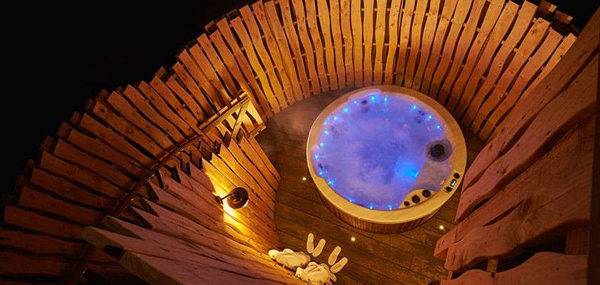 Circular hot tub bubbles with blue LED lights, steaming water swirling. Surrounded by curved wooden slat walls and deck at night, warm lamp glow, towels and slippers placed nearby.