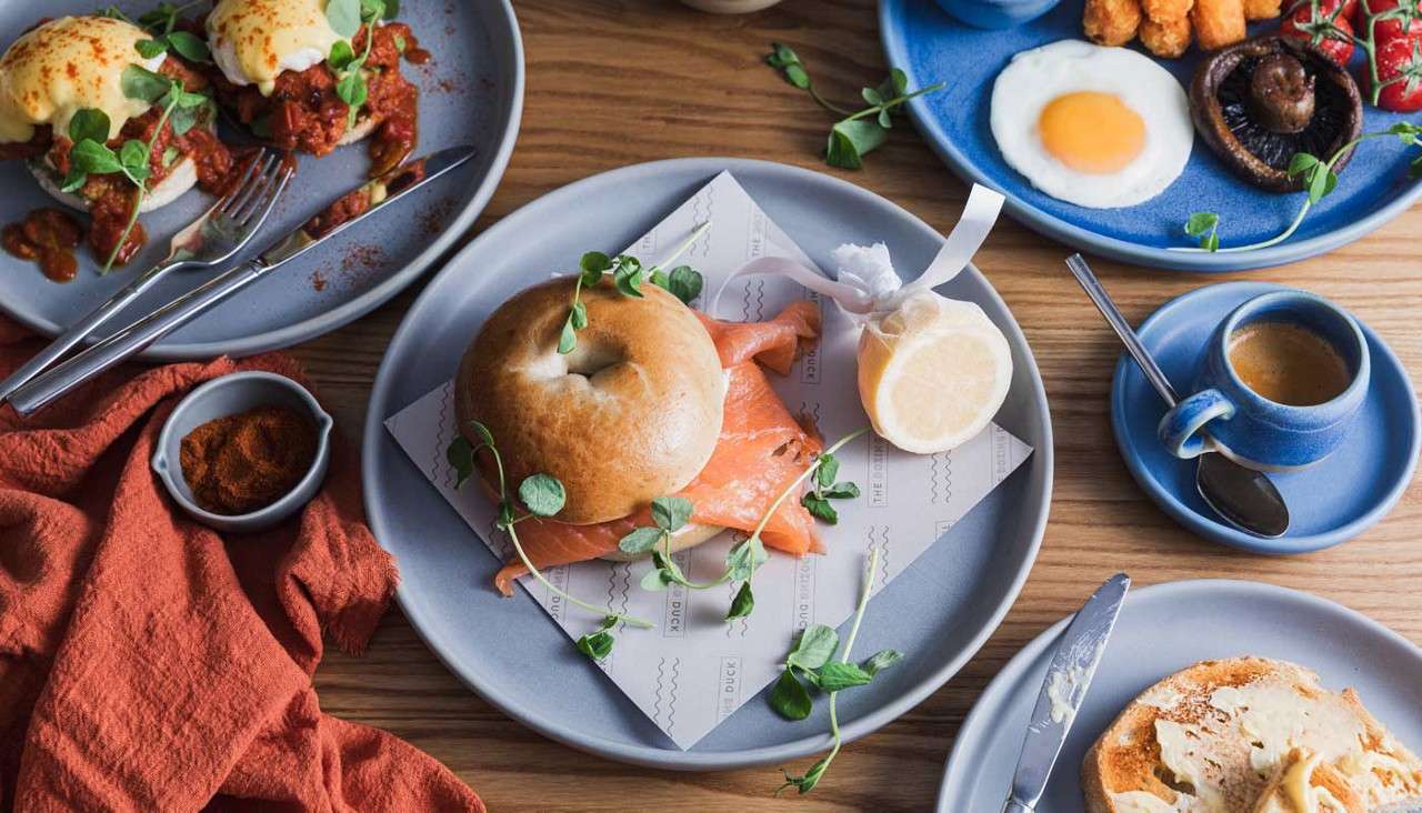 Bagel sandwich sits on a plate with smoked salmon and greens; around it, breakfast dishes include fried egg, mushroom, tomatoes, hash browns, eggs Benedict, toast, and espresso on a wooden table.