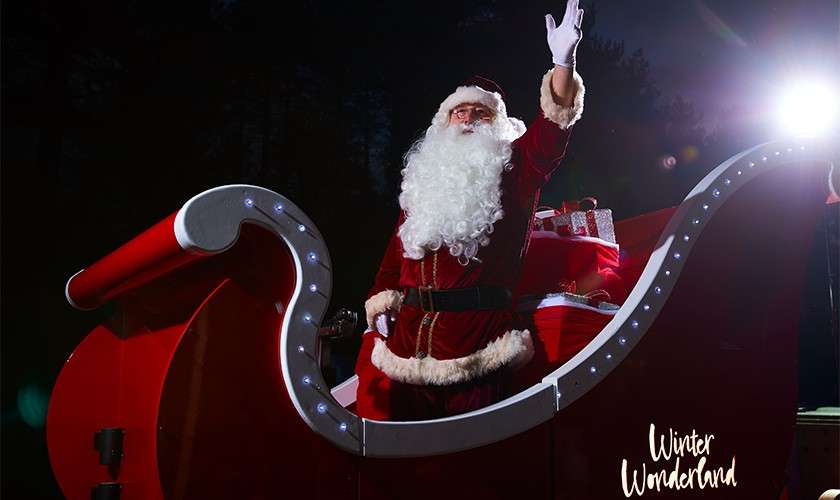 Person dressed as Santa Claus waves while standing in an illuminated red sleigh; wrapped gifts behind. Nighttime outdoor scene with spotlight and dark trees. Winter Wonderland