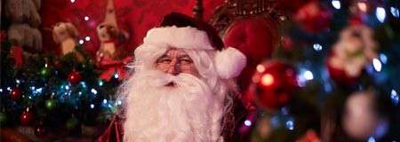 Person dressed as Santa Claus sits smiling, partially obscured by colorful Christmas tree ornaments and lights, in a warmly lit, red-decorated room with festive garlands and figures in the background.