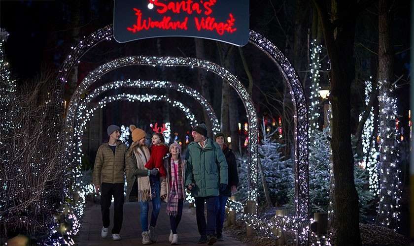 Group of five adults and one child walk, chatting, under glowing archways on a forest path at night; trees sparkle with white lights. Text overhead: Santa's Wonderland Village.