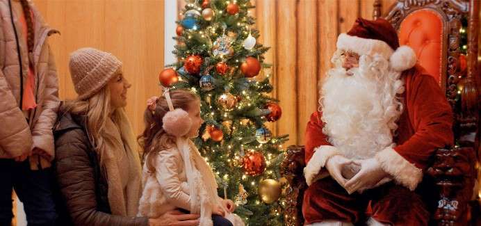 Person dressed as Santa Claus sits on an ornate chair, facing a child beside a kneeling adult. They interact near a lit, ornamented Christmas tree in a warm indoor setting.