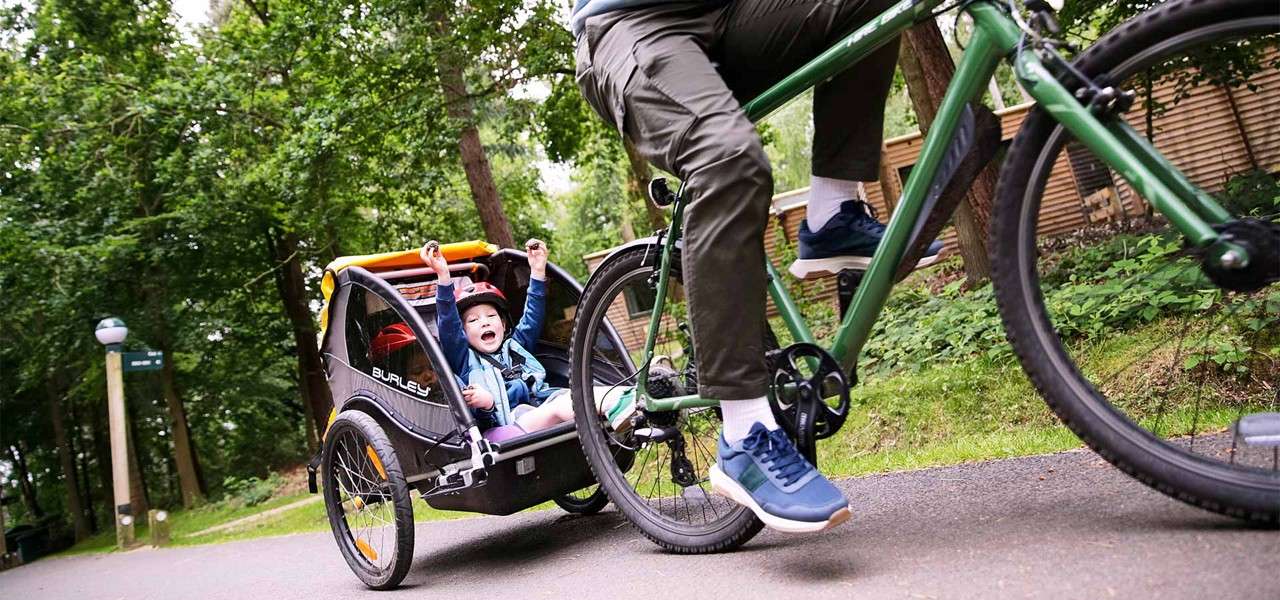Child in a bike trailer cheers with arms raised, while an adult pedals a green bicycle towing it along a paved park path amid trees and fencing. Text: BURLEY.