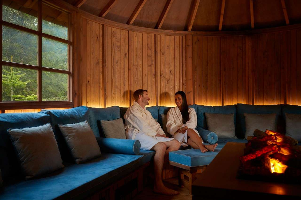 Two people in white bathrobes sit chatting on blue-cushioned benches, relaxing beside a glowing fire pit inside a circular wooden lounge, with a window showing green trees outside.