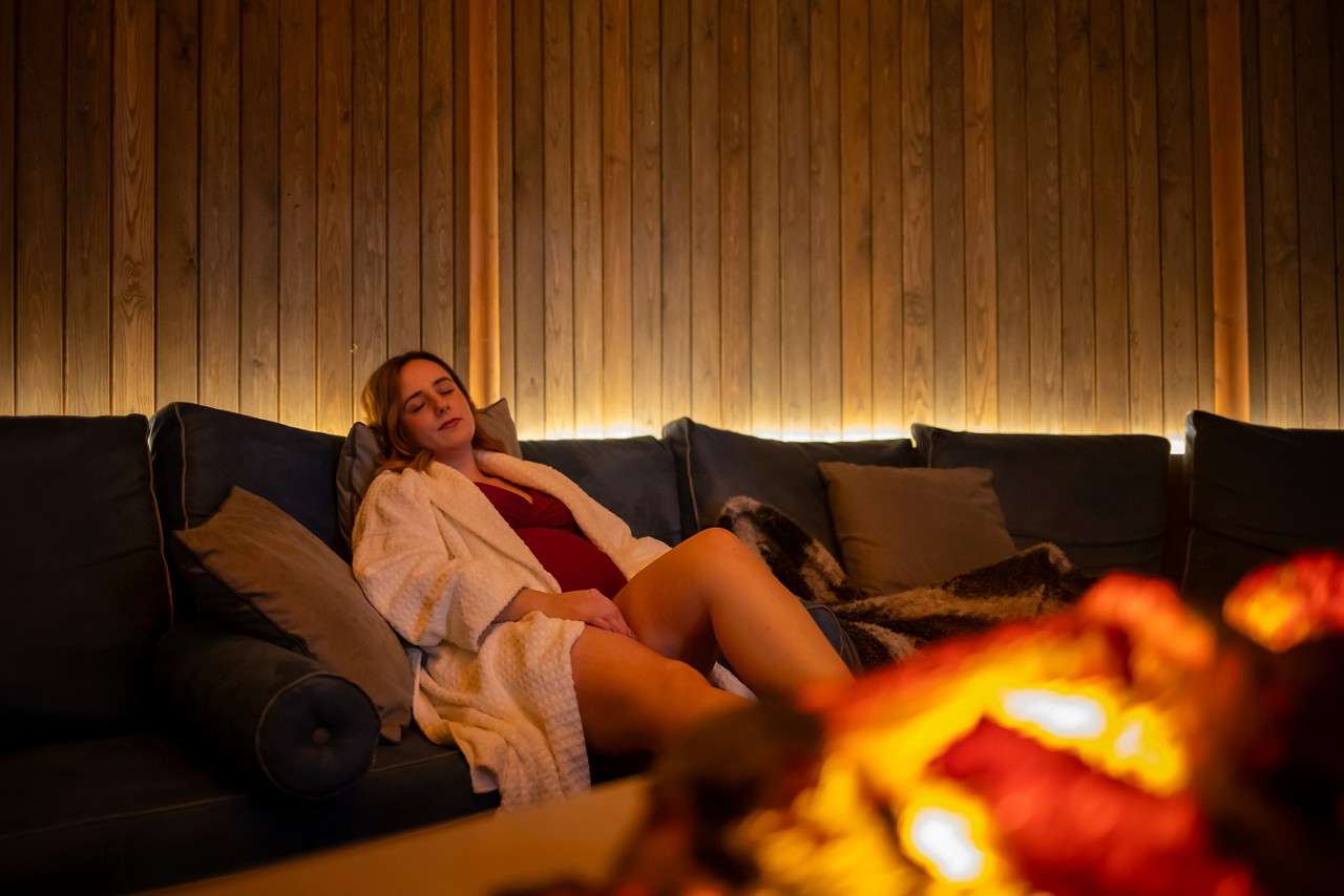 Person in a white robe reclines with eyes closed on a cushioned sofa, relaxing. Warm ambient lighting glows behind wood-paneled wall; soft blanket and pillows nearby; fireplace blur in foreground.