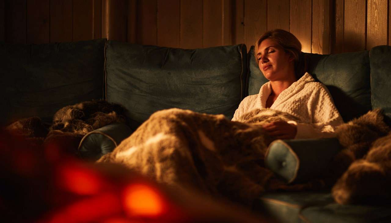 Woman relaxes, eyes closed, wrapped in a thick blanket on a blue sofa; warm orange firelight glows in the foreground; wood-paneled walls and plush cushions create a cozy indoor setting.