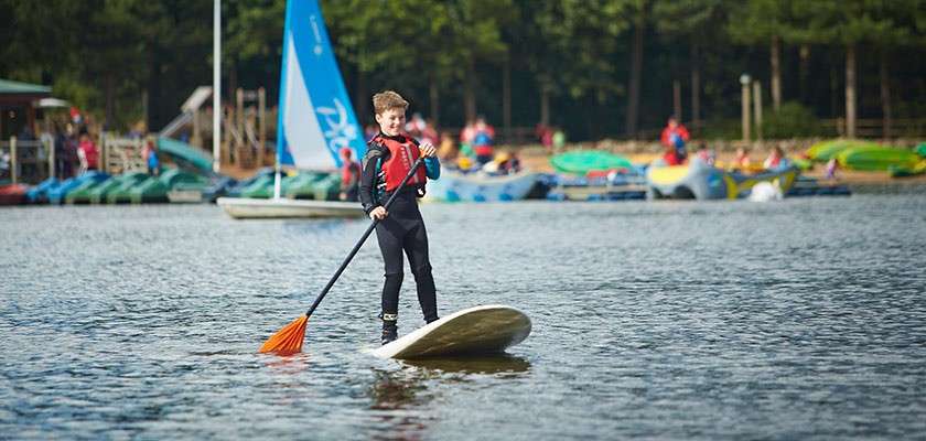 Child on a stand-up paddleboard paddles forward, balancing on calm lake water. Behind, a watersports center shows kayaks, a blue sailboat, and people near trees. Visible text: “Pico” on the sail.