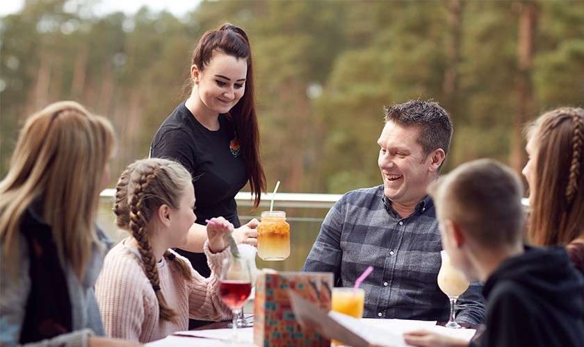 Server hands a mason-jar iced drink to a smiling diner while others watch. Seated group holds menus and beverages on a table, on an outdoor patio overlooking trees.