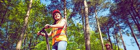 Rider operates a handlebar electric scooter/Segway, wearing a red helmet and orange safety vest, gliding along a forest trail; tall trees surround them, with another rider visible in the background.