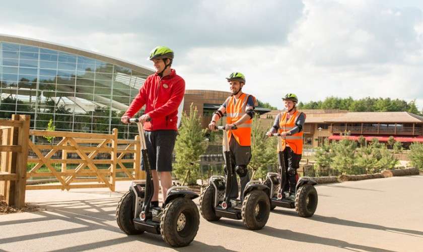 Three people on Segways ride in a line, wearing helmets and safety vests, along a paved path in a landscaped park beside modern glass and wooden buildings under cloudy sky.