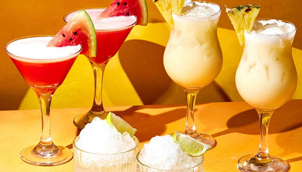 Colorful cocktails sit on a table: red martinis with watermelon wedges, creamy yellow drinks with pineapple slices, and two short glasses of crushed ice with lime, against a yellow backdrop.