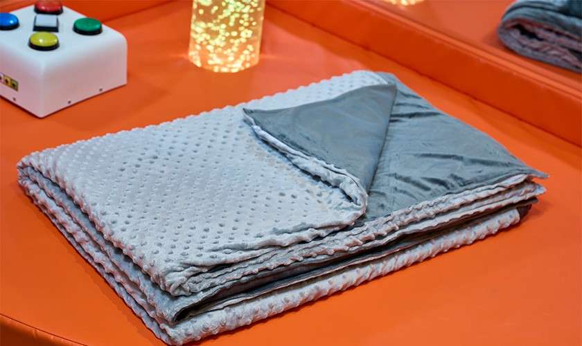 Folded gray textured blanket lies with a corner flipped, revealing smooth lining, on an orange padded surface beside a white box with colored buttons and a glowing bubble tube.