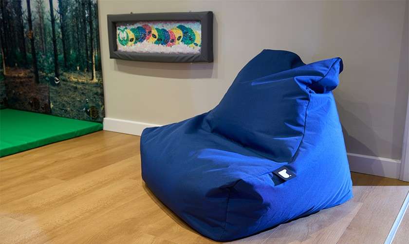 Blue beanbag chair rests on a wooden floor, angled toward a wall. Nearby, a green floor mat and a framed sensory panel hang beneath a forest-themed wall mural.