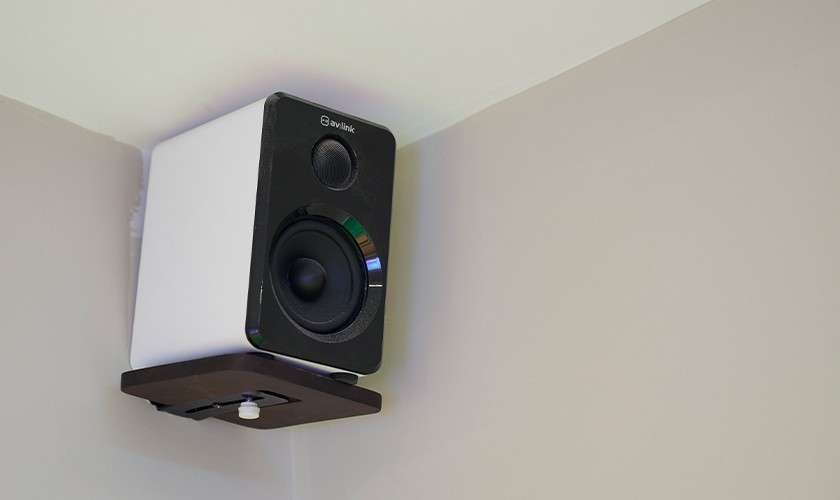 Speaker tilts downward on a small wall bracket in a ceiling corner, white cabinet with black front. Text visible: av:link.