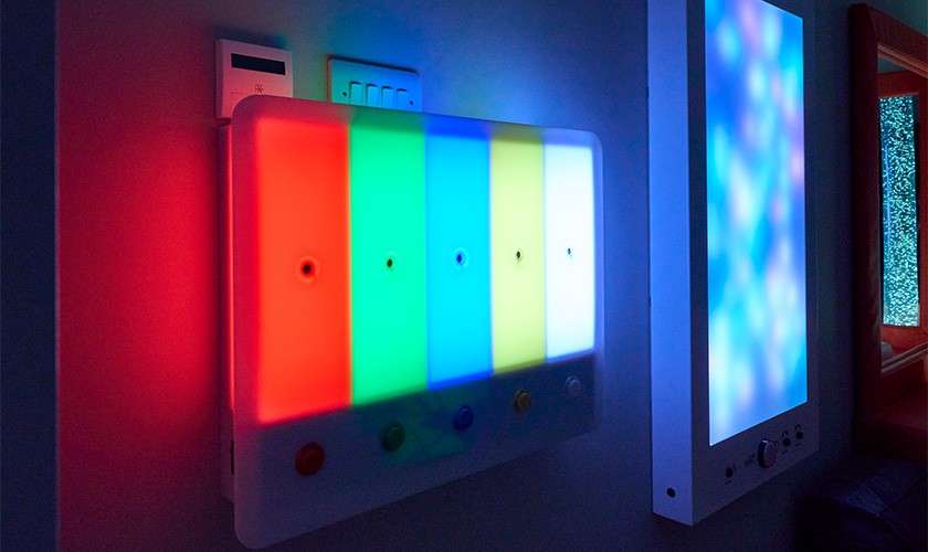 Wall-mounted LED panel glows in vertical red, green, blue, and yellow bars, with small buttons below; beside it, another soft-color light panel shimmers, illuminating a dim, modern interior hallway.