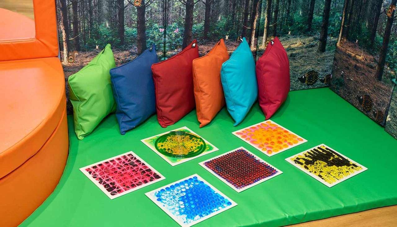 Colorful cushions line a wall, resting upright; six textured sensory tiles lie arranged on a green padded floor in a children’s play or sensory corner with a forest-themed backdrop.