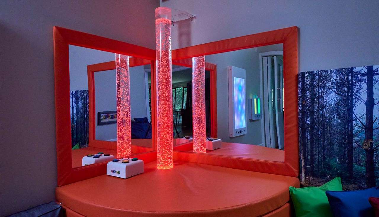 Bubble tubes glow red, bubbles rising; mirrors reflect them. In a sensory room: orange-padded base and frames, cushions, wall light panels, and control boxes with colored buttons labeled “TFH.”