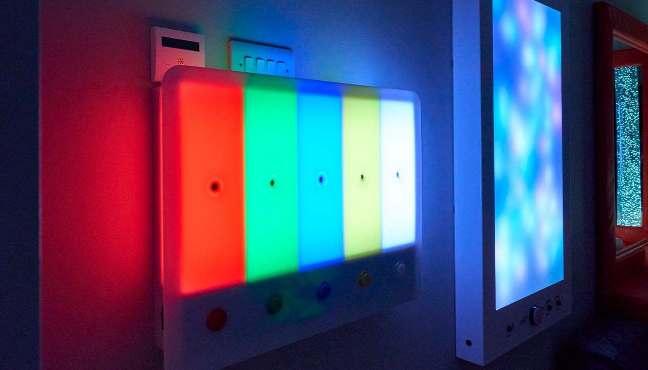 Multicolored LED wall panel glows in vertical red, green, blue, cyan, yellow, and white bars, with buttons below, mounted in a dim room beside another softly lit panel.