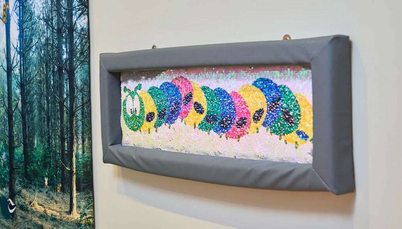 Framed sequin artwork depicts a colorful cartoon caterpillar moving left; mounted on a white wall next to a large photograph of a pine forest.