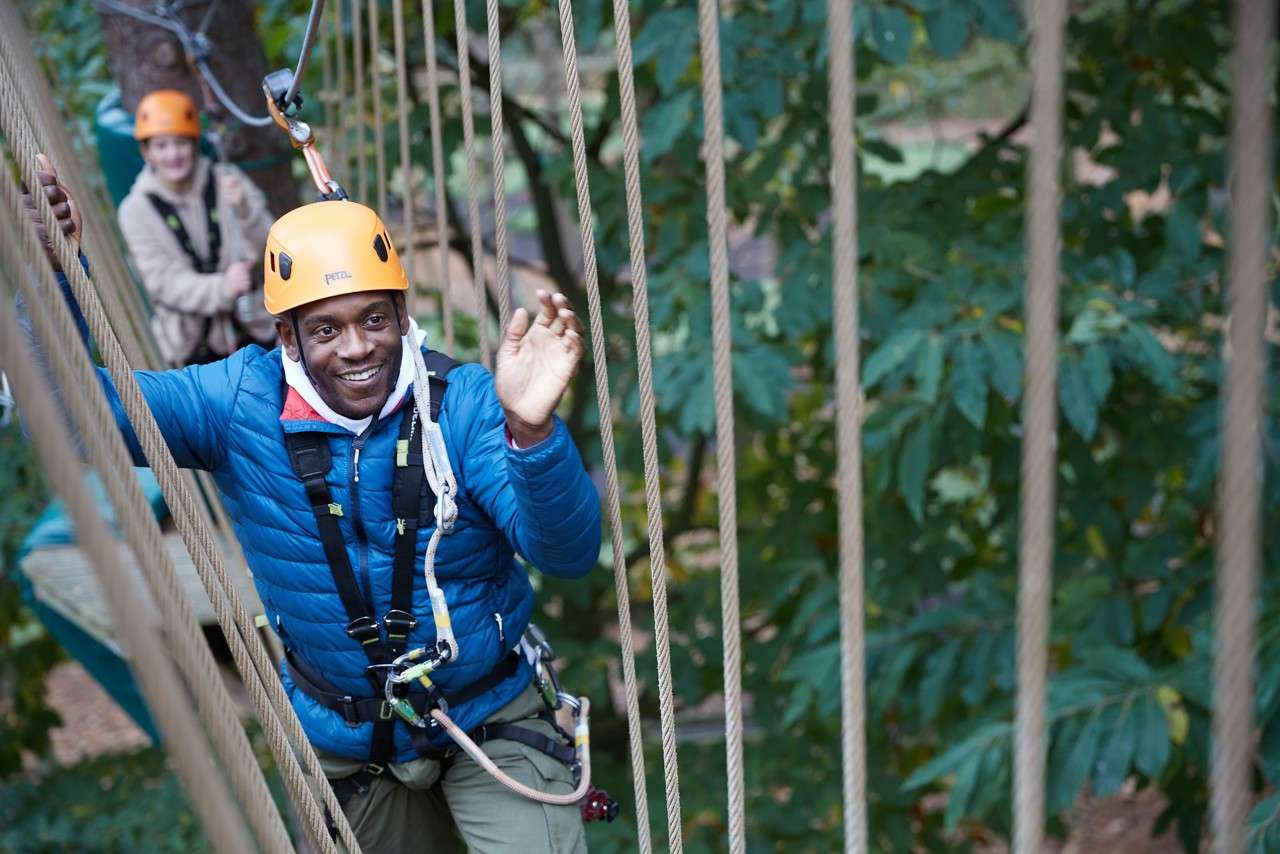 Man wearing orange helmet waves while balancing on rope bridge; clipped to harness and carabiners; another participant follows; forest canopy surrounds. Text: "PETZL" on helmet.