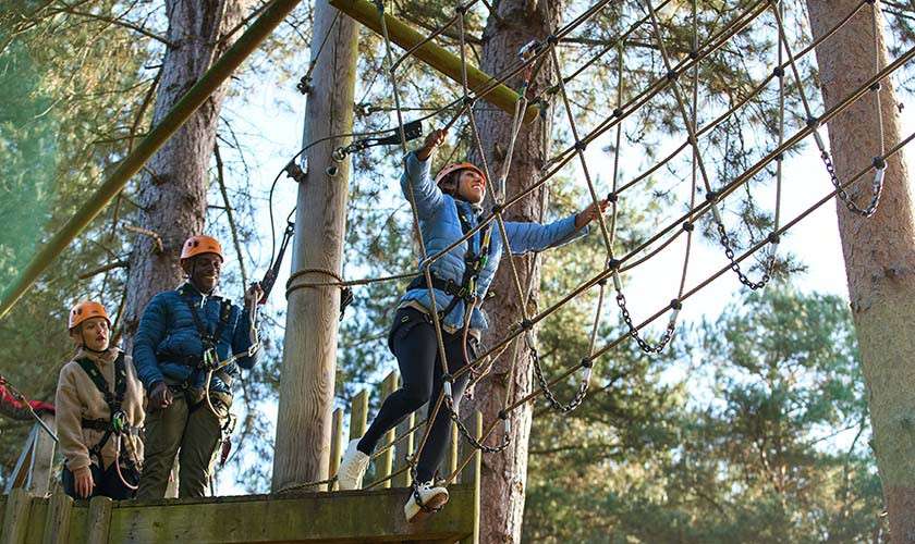 A helmeted climber steps across a high-ropes net, balancing with arms out. Two harnessed teammates watch from a wooden platform. Tall pine trees surround the elevated obstacle course in daylight.