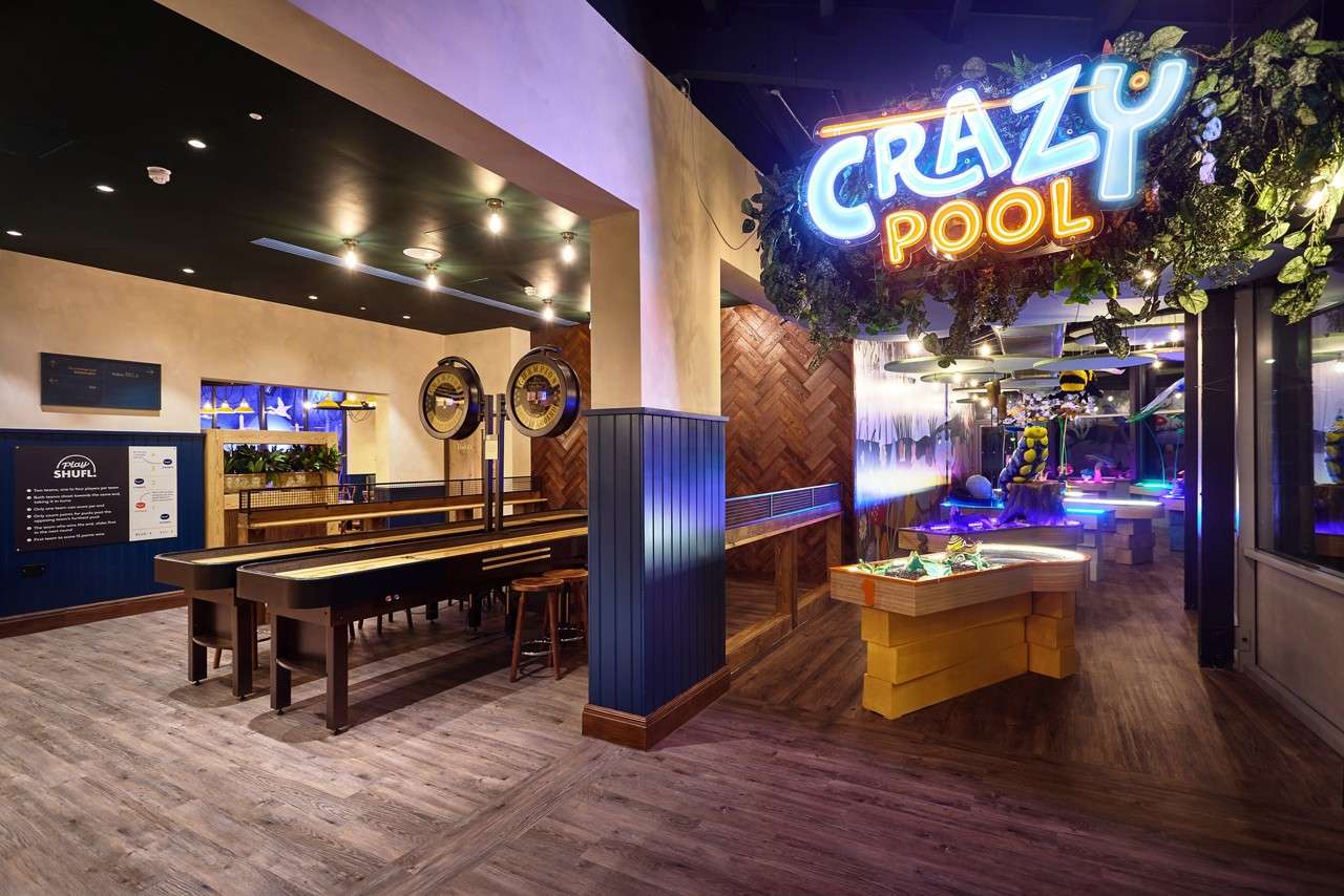 Neon sign “CRAZY POOL” glows above playful gaming area; adjacent shuffleboard lanes stand empty under warm lights in a modern bar with wood floors, plants, and arcade-style tables.